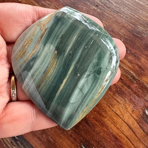 May include: A polished, gemstone in shades of green, tan, and white. The stone has a smooth, rounded shape and a glossy finish. The natural stone is held in a hand, showcasing its unique patterns and colors. The gemstone is a decorative item.