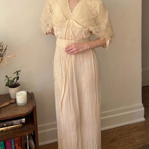 May include: A vintage cream-colored dress with lace detailing. The dress features a V-neckline, short sleeves, and a flowing skirt. The bodice has a satin-like texture, and the sleeves and neckline are adorned with delicate lace. The dress appears to be made of a lightweight fabric.