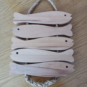 May include: A wooden wall hanging with five fish-shaped elements. Each fish has a small, dark eye and a tail fin. The fish are joined vertically and suspended by a rope loop. The wood has a natural, light brown colour.