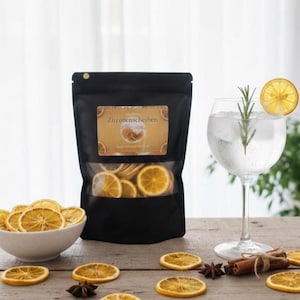 Puede incluir: Una bolsa negra de rodajas de naranja deshidratadas con el texto "Zitronenscheiben" en la etiqueta. Una copa de cóctel con una rodaja de limón y una guarnición de romero está al lado de la bolsa. Un cuenco de rodajas de naranja y otras guarniciones están sobre una superficie de madera.