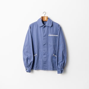 May include: A blue work jacket with a collar, long sleeves, and buttoned cuffs. The jacket features a white zip on the chest pocket and two side pockets. It hangs on a wooden hanger against a white backdrop.