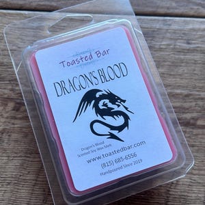 Puede incluir: Un derretimiento de cera rosa en un recipiente de plástico transparente con una etiqueta que dice "Toasted Bar Dragon's Blood Scented Soy Wax Melt www.toastedbar.com (815) 685-6556 Handpoured Since 2019". La etiqueta también presenta una ilustración en blanco y negro de un dragón.