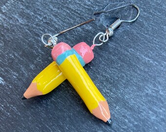 May include: A pair of yellow pencil earrings with pink and blue eraser details. The earrings are made of polymer clay and have silver hooks.