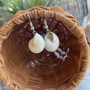 May include: A pair of white seashell earrings with silver hooks. The earrings are hanging from a wicker basket.