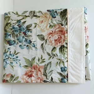 May include: A folded pillowcase featuring a floral pattern with large peach and cream-colored peonies and smaller blue flowers on a white background. The side panel is a solid cream color, separated from the floral print by a thin, light-colored piping.