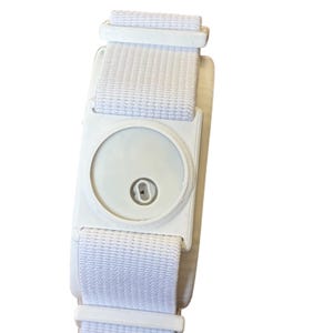 May include: A white wristband featuring a circular, light-coloured device at its centre. The band is made of a woven material and has a buckle closure. The device has a small, dark-coloured centre.