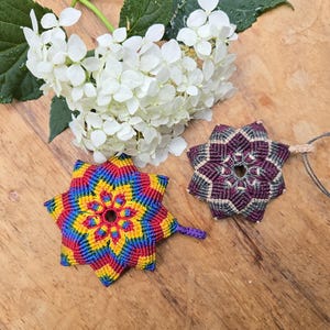 Peut inclure: Deux ornements en macramé en forme d'étoile faits à la main, aux couleurs vives, un arc-en-ciel et un bordeaux, sont posés sur une surface en bois. Une grappe de fleurs d'hortensia blanches avec des feuilles vertes est également présente.