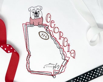 May include: A white fabric garment featuring an embroidered outline of the state of Georgia in red and black thread. A bulldog sits atop the state shape, which includes a brown football icon and the word Georgia in red, cursive-style lettering.