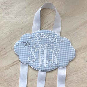 May include: A blue and white gingham ribbon holder with a white embroidered monogram "S.M.K." and a bow. The holder has three white ribbons attached.