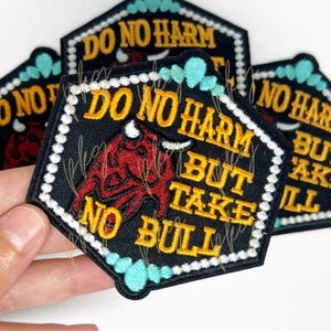 May include: Black embroidered patch with the text "DO NO HARM BUT TAKE NO BULL" in yellow. The patch features a red bull illustration and turquoise accents. The patch has a white border.
