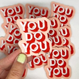 Puede incluir: Parches bordados con la frase "YOU DO YOU" en rojo y blanco. Los parches tienen un fondo rosa claro. Una mano con una uña pintada de amarillo sostiene algunos de los parches.