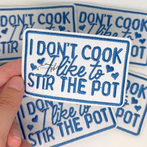 May include: A rectangular embroidered patch with a white background and a blue border. The text reads I DON'T COOK I like to STIR THE POT in blue thread, accented with small blue embroidered hearts scattered around the lettering.