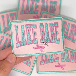 May include: A rectangular embroidered patch with a light pink background and a teal border. The text "LAKE BABE" is stitched in teal with a pink outline. Below the text is a small pink embroidered icon of two crossed oars.