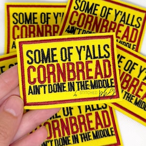 Puede incluir: Un parche rectangular bordado con fondo amarillo y borde rojo. El texto en negro y rojo dice: SOME OF Y'ALLS CORNBREAD AIN'T DONE IN THE MIDDLE. El parche presenta un patrón de costura denso y texturizado en todo el diseño.