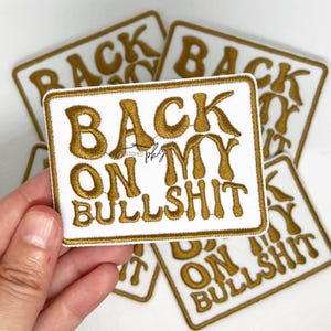 May include: Gold embroidered iron-on patch with the text "Back on my Bullshit" on a white background.