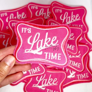 May include: A collection of pink embroidered patches, each with the white script text "IT'S Lake Time". The patches have a red and white border. The image shows a textured fabric.