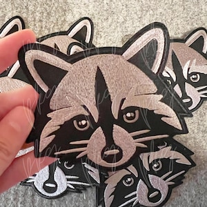 Puede incluir: Un conjunto de cinco parches bordados para planchar con la cara de un mapache en negro y gris con detalles blancos.