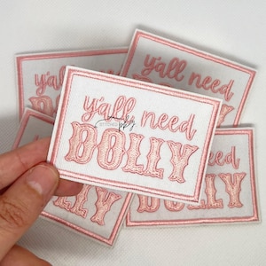 Może przedstawiać: Prostokątne białe naszywki z różowym haftowanym tekstem "y'all need DOLLY". Naszywki mają różową obwódkę i są ułożone jeden na drugim. Słowo "STITCHED" jest również wyhaftowane na naszywce.