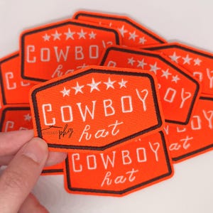 May include: Orange embroidered patch with the words "COWBOY hat" in white block letters. The patch has a black border and five white stars at the top. Several patches are visible.