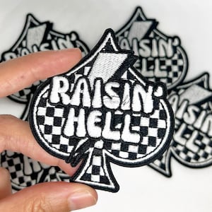 Op de afbeelding: Zwart en wit geborduurde patch in de vorm van een schoppen. De patch heeft de woorden "RAISIN' HELL" in het wit, met een geblokt patroon in het midden. De patch is omgeven door een zwarte rand.