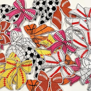 May include: A selection of embroidered bow patches with sports themes. Designs include soccer balls, American footballs, basketballs, and baseballs. The bows are in pink, white, orange, yellow, and black. Perfect for crafting and adding a sporty touch to clothing and accessories.