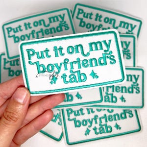 Pode incluir: Patch retangular branco com texto bordado turquesa que diz "Put it on my boyfriend's tab". O patch tem uma borda turquesa. Vários patches semelhantes estão no fundo. O patch é segurado em uma mão.