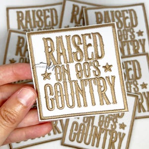 Op de afbeelding: Witte vierkante patch met goud geborduurde tekst "RAISED ON 90'S COUNTRY". De patch heeft een gouden rand en steraccenten. Op de achtergrond zijn verschillende vergelijkbare patches zichtbaar.