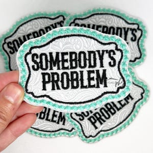 May include: Four white embroidered iron-on patches with a teal border. The patches feature the text "Somebody's Problem" in black lettering.