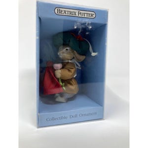 May include: A Beatrix Potter collectible doll ornament inside a blue box. The rabbit figure wears a dark green hat, a tan coat, and holds a red fabric accessory with a small pink flower. The box features the text Beatrix Potter and Collectible Doll Ornament.
