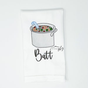 May include: White kitchen towel with black embroidery of a pot of soup with a spoon and the text "Batt" below. The text "Stitched by" is also embroidered in black.