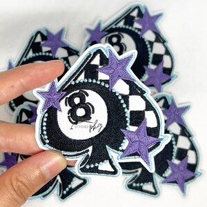 May include: Embroidered patch in the shape of a black spade with a white and black checkered design. The patch features a white eight ball design with the number 8. Purple star accents are scattered around the patch.
