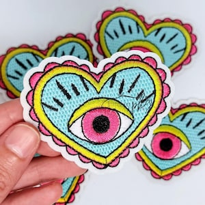 May include: An embroidered heart-shaped patch featuring a light blue center with a central pink eye, a black pupil, and black eyelashes. The heart is outlined in yellow and finished with a scalloped pink border. The text Stitched Peggy is visible.