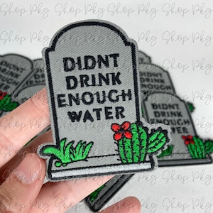 May include: A gray tombstone-shaped patch with green grass and a red cactus flower. The text on the patch reads "Didn't Drink Enough Water".