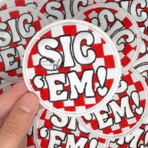 Op de afbeelding: Ronde geborduurde patch met de tekst "SIC EM!" in wit, omlijnd met zwart, op een rood-witte geblokte achtergrond. De patch heeft een witte rand. Meerdere patches zijn zichtbaar.