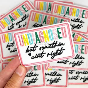 Könnte beinhalten: Gestickter Aufnäher mit den Worten "UNDIAGNOSED but somethin ain't right" in farbenfrohen Buchstaben. Der Aufnäher hat einen weißen Hintergrund mit einem rosa Rand. Mehrere Aufnäher sind sichtbar.
