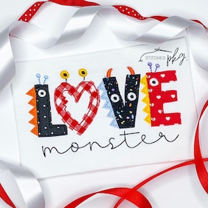 May include: A white fabric with the words "LOVE monster" embroidered in black thread. The letters are decorated with colorful felt and have googly eyes. The "O" in LOVE is a red heart with a white and red checkered pattern.