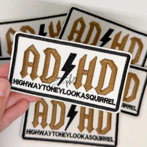 May include: A rectangular embroidered patch with a white background and a black border. The patch features the gold-coloured text ADHD with a black lightning bolt in the centre, and the text HIGHWAYTOHEYLOOKASQUIRREL in black below it.