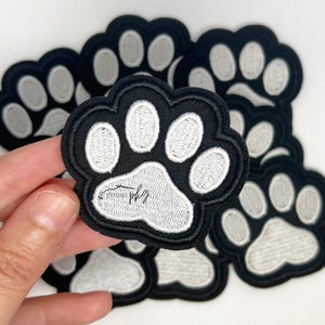 May include: A set of black and white paw print iron-on patches. The patches are embroidered with white paw prints on a black background.