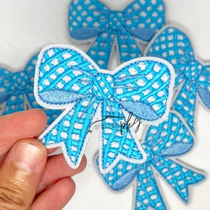 May include: Embroidered blue bow applique with a white dotted pattern and a white border. The bow is held in a hand, with several other bows visible in the background. The bows are likely for crafting or embellishment.