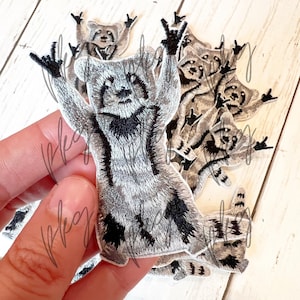 May include: A set of 10 iron-on patches featuring a raccoon with its arms raised in a rock and roll hand gesture. The patches are grey and black with embroidered details.