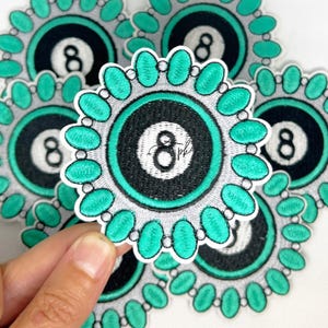 May include: Embroidered patch featuring a black and white eight ball design with a teal border and accents. The patch has a white border and a floral-like design around the edge. The number 8 is prominently displayed in the center.