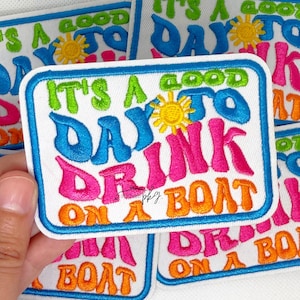 May include: A rectangular embroidered patch with a white background and a blue border. The text reads IT'S A GOOD DAY TO DRINK ON A BOAT in lime green, blue, pink, and orange thread. A yellow sun icon is positioned above the word DRINK.