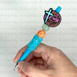 May include: A blue pen with a turquoise and clear glitter design. The pen has a pink and orange bead with a white bead and a charm with the words "Pray on it, Pray over it, Pray through it" and a cross.