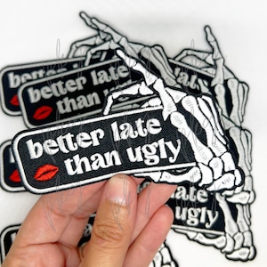 Puede incluir: Parche bordado en blanco y negro con la frase "better late than ugly" y un diseño de labios rojos. El parche rectangular con esquinas redondeadas presenta una mano de esqueleto blanca.