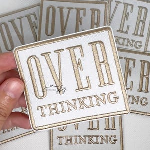 以下が含まれることがあります： 「OVER THINKING」と金色の刺繍が施された白い正方形のパッチ。 パッチには金の縁取りがあり、他の同様のパッチと一緒に表示されます。 パッチはおそらく衣類やアクセサリー用です。