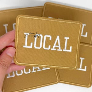 May include: A rectangular mustard yellow embroidered patch with a stitched border. The word LOCAL is embroidered in white capital letters across the centre. A small black script logo is visible above the letter O in the word LOCAL.