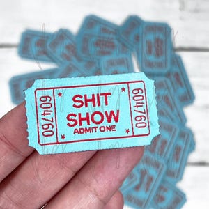 Puede incluir: Un boleto azul claro con texto rojo que dice "SHIT SHOW" y "ADMIT ONE". El número del boleto es 609/760.