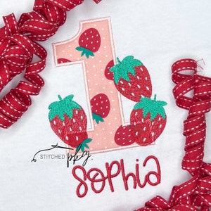 May include: A pink and white embroidered number one with strawberries. The number one is surrounded by strawberries. The text "Stitched pky. Sophia" is embroidered below the number one.