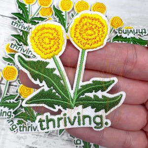May include: A white embroidered patch with two yellow dandelions and green leaves. The patch has the word "thriving" written in green below the dandelions.