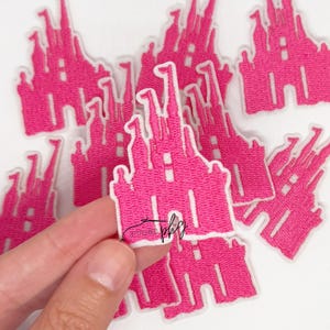 May include: A collection of pink embroidered castle patches with white borders. The patches feature a detailed castle design with turrets and windows. The patches are held in a hand, with several more scattered in the background.